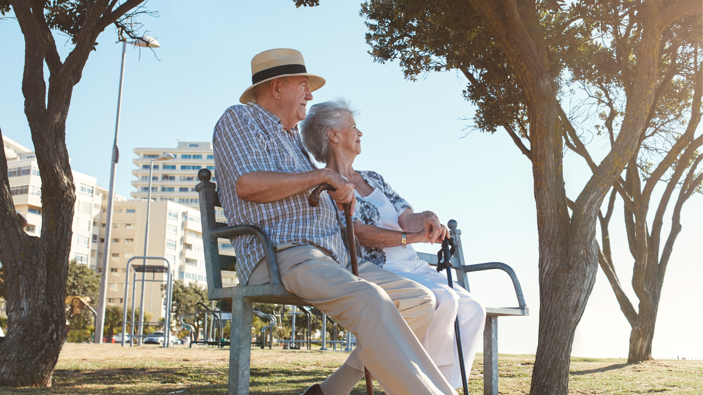 Elderly couple sitting on a bench near trees and residential buildings in a South African retirement community.